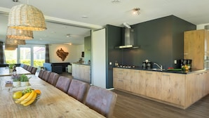 A modern kitchen with wooden cabinets, a large island, and a dining area with a long table and leather chairs. - Countryside Group Getaway (Paasloo)