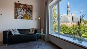 A modern living room with a black sofa, a glass coffee table, and a large window with a view of a church tower. - Apartment in Koudekerke Near Seabeach (Koudekerke)