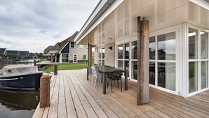 A wooden deck with a table and chairs, a boat docked by the water, and a house with large windows and a brick facade. - Villa With a Sauna in Tjeukemeer (Delfstrahuizen)