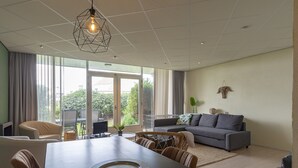 A modern living room with a dining table, sofa, and large windows offering a view of greenery. - Nice Apartment With Garden Near the North Sea (Julianadorp)