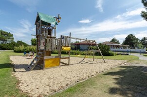 Un parque infantil con una estructura de madera, que incluye un tobogán y una zona de escalada, ubicado en un área de arena con césped y árboles circundantes.