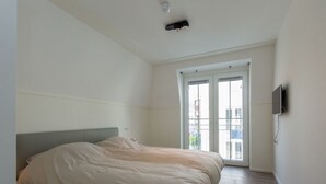 A bedroom with a bed, a TV mounted on the wall, and a window with a view of buildings.