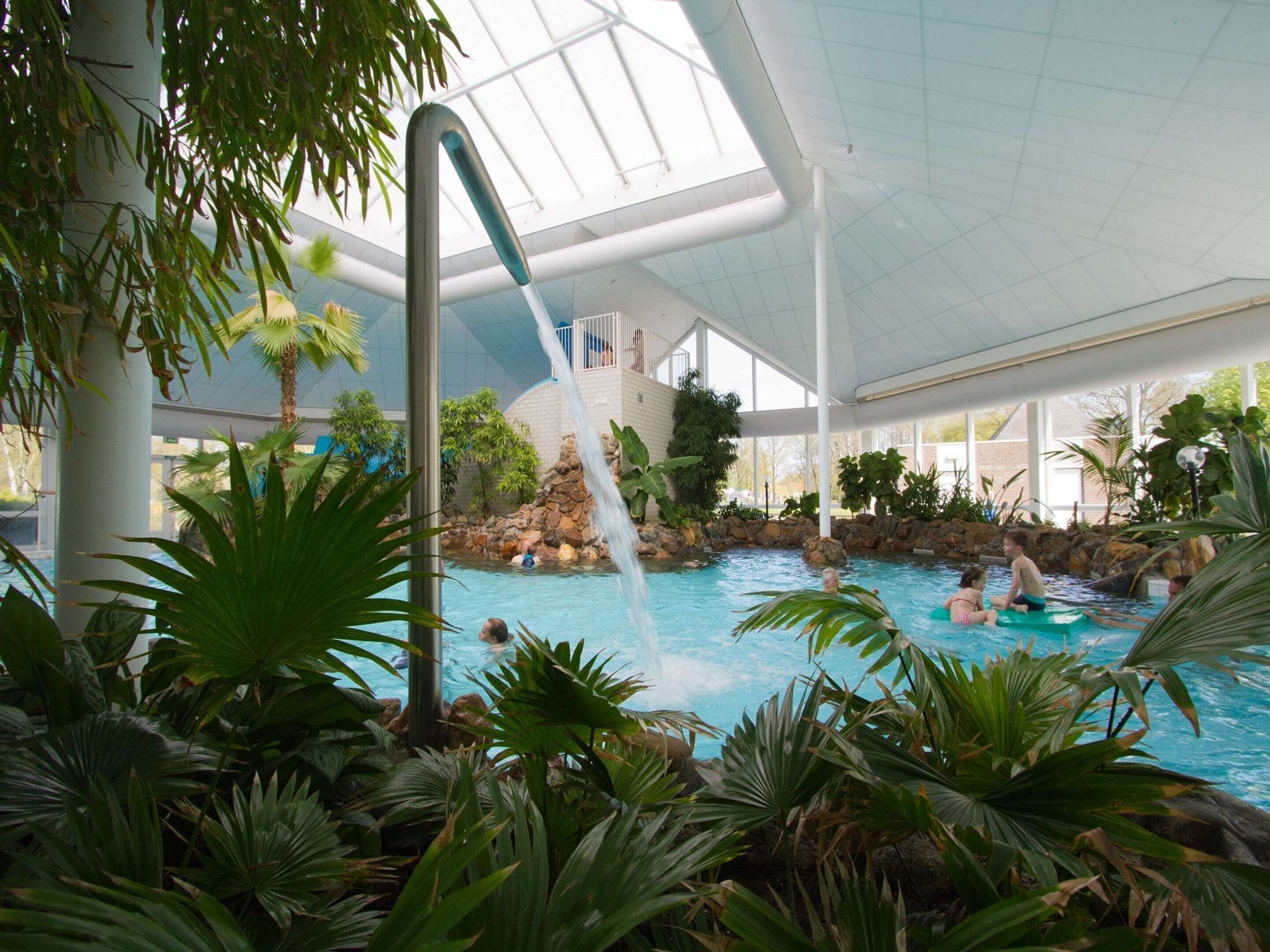 An indoor swimming pool with a waterfall feature, surrounded by tropical plants and a glass ceiling.