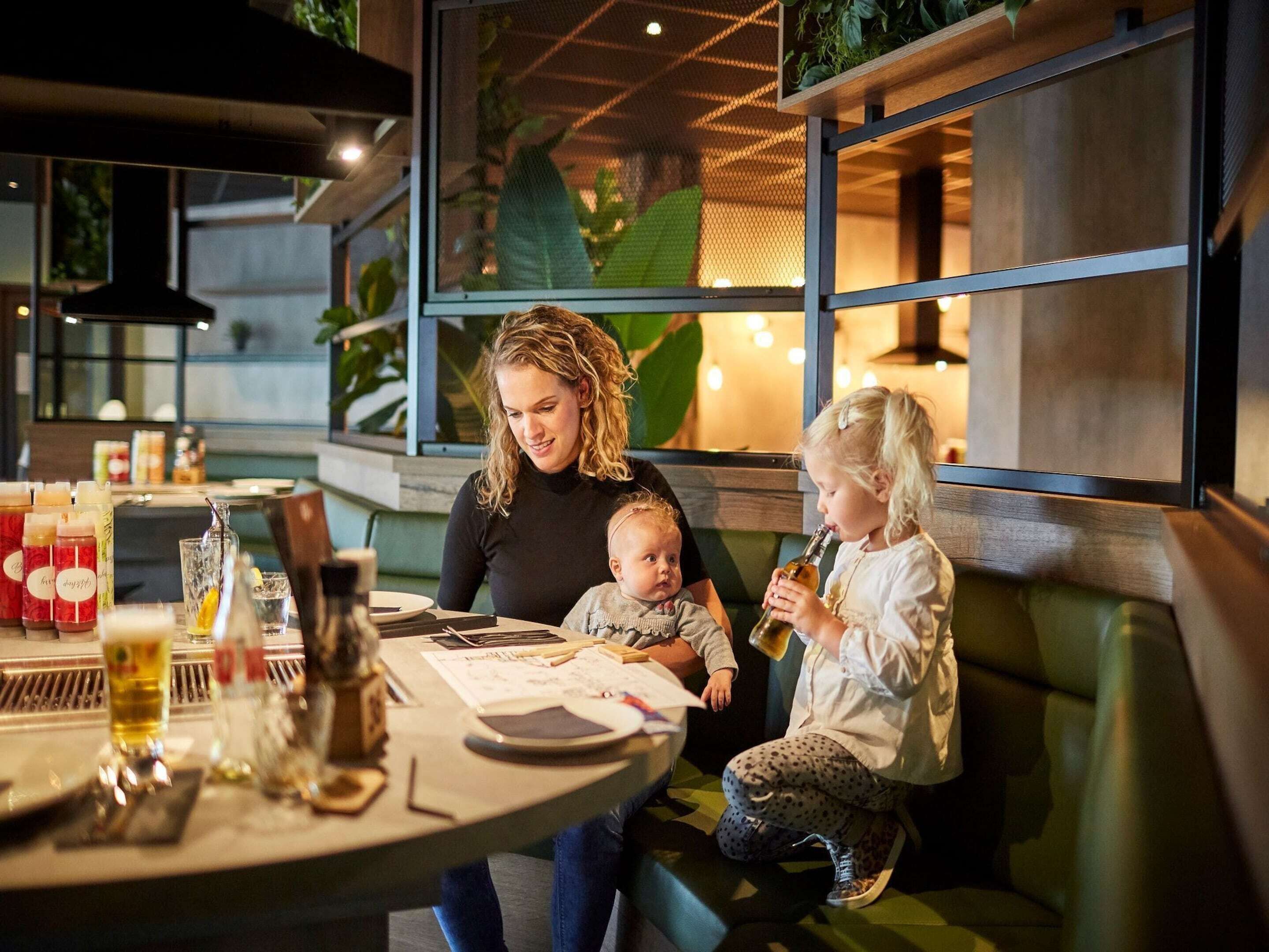 A woman and two children at a table in a restaurant with a modern interior, including a green couch and a window with plants.