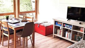 Ein Zimmer mit einem Holzesstisch, Stühlen, einem Bücherregal, einem Fernseher und großen Fenstern mit Blick auf Grünflächen.