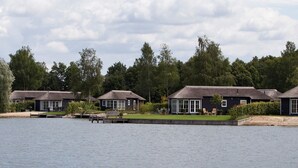 A row of dark-colored cottages with thatched roofs, situated by a calm lake, surrounded by lush greenery. - Cosy Holiday Home in Twente (Hoge Hexel)