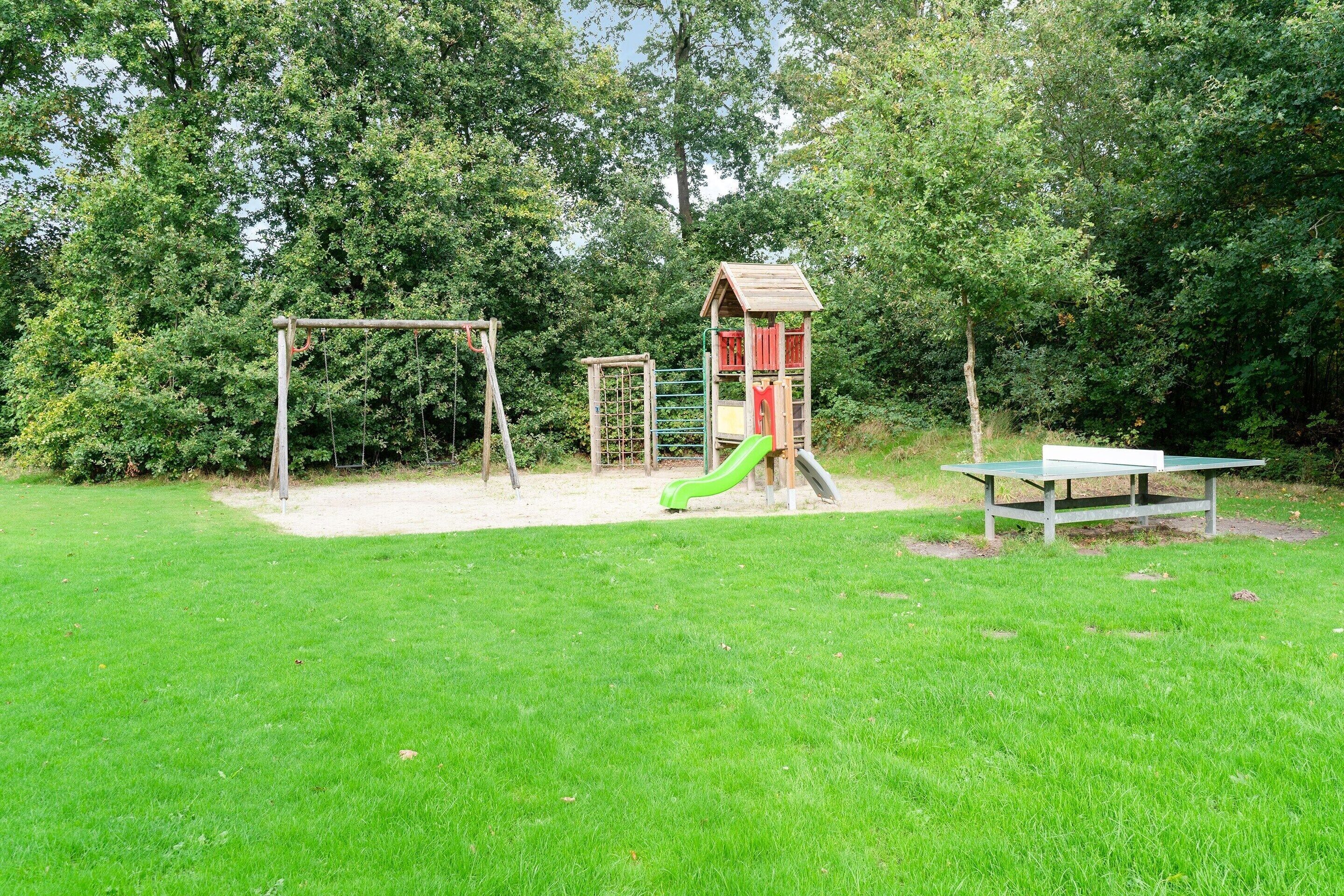A playground with a swing set, slide, and climbing structure surrounded by grass and trees.
