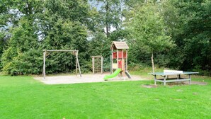 Ein Spielplatz mit Schaukel, Rutsche und Klettergerüst, umgeben von Gras und Bäumen.
