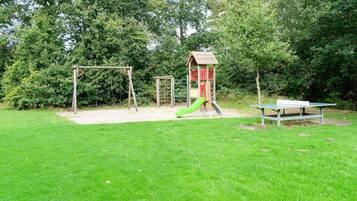 A playground with a swing set, slide, and climbing structure surrounded by grass and trees.