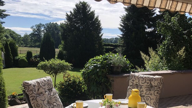 An outdoor patio with a table set for two, surrounded by wicker chairs, under a covered area with a view of a green landscape. - Vacation apartment "Zum Wiesenblick" in the heath village of Eimke, 130 sqm  (Eimke)