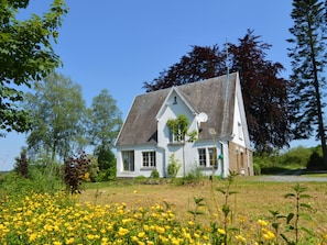 A white house with a steep roof, surrounded by a field of yellow flowers and trees. - A Superb Cottage in the Middle of a Natural Park (Hotton)