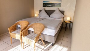 Un dormitorio con una cama, dos sillones de mimbre, una mesita, una mesilla de noche y un cuadro en la pared.