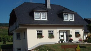 Una casa de dos pisos con techo oscuro, ventanas blancas y fachada color beige. Hay macetas con flores en el porche delantero y un muro de piedra al frente.