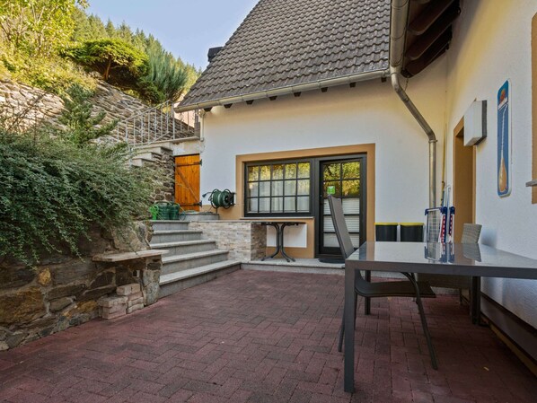 Una casa con escaleras de piedra, un patio con mesa y sillas, y una ventana que ofrece vistas del entorno verde.