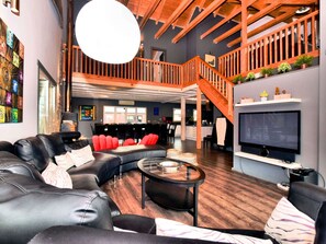 A spacious living area with a large leather sofa, a coffee table, and a TV mounted on the wall. A wooden staircase leads to an upper level, and there are potted plants on a shelf. - Beautiful Villa with Heated Outdoor Pool, Sauna, Hot Tub in Forest (Manhay)