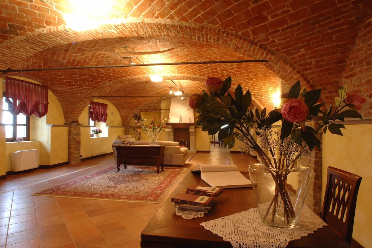 Una habitación amplia con un alto techo abovedado de ladrillo, un gran ventanal con cortinas rojas, una mesa de madera con libros y un jarrón de flores, y una zona de estar acogedora con un sofá y una lámpara.