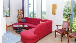 A living room with a red sofa, a wooden chair, a glass coffee table, and a view of greenery through large windows. - Villa mit Sauna und Garten (Schotten)