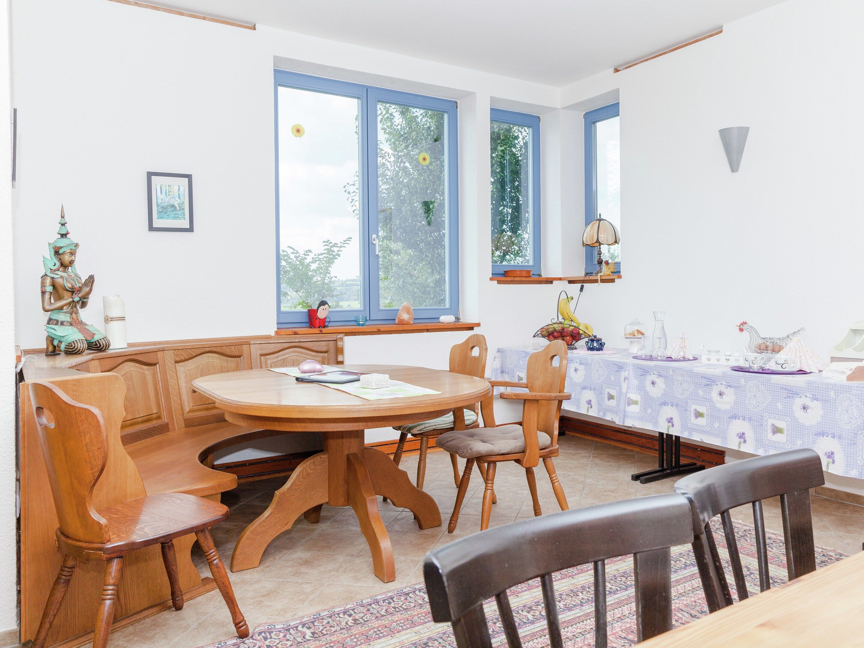A dining area with a round wooden table, four chairs, a window with blue frames, and a wall-mounted shelf with decorative items.