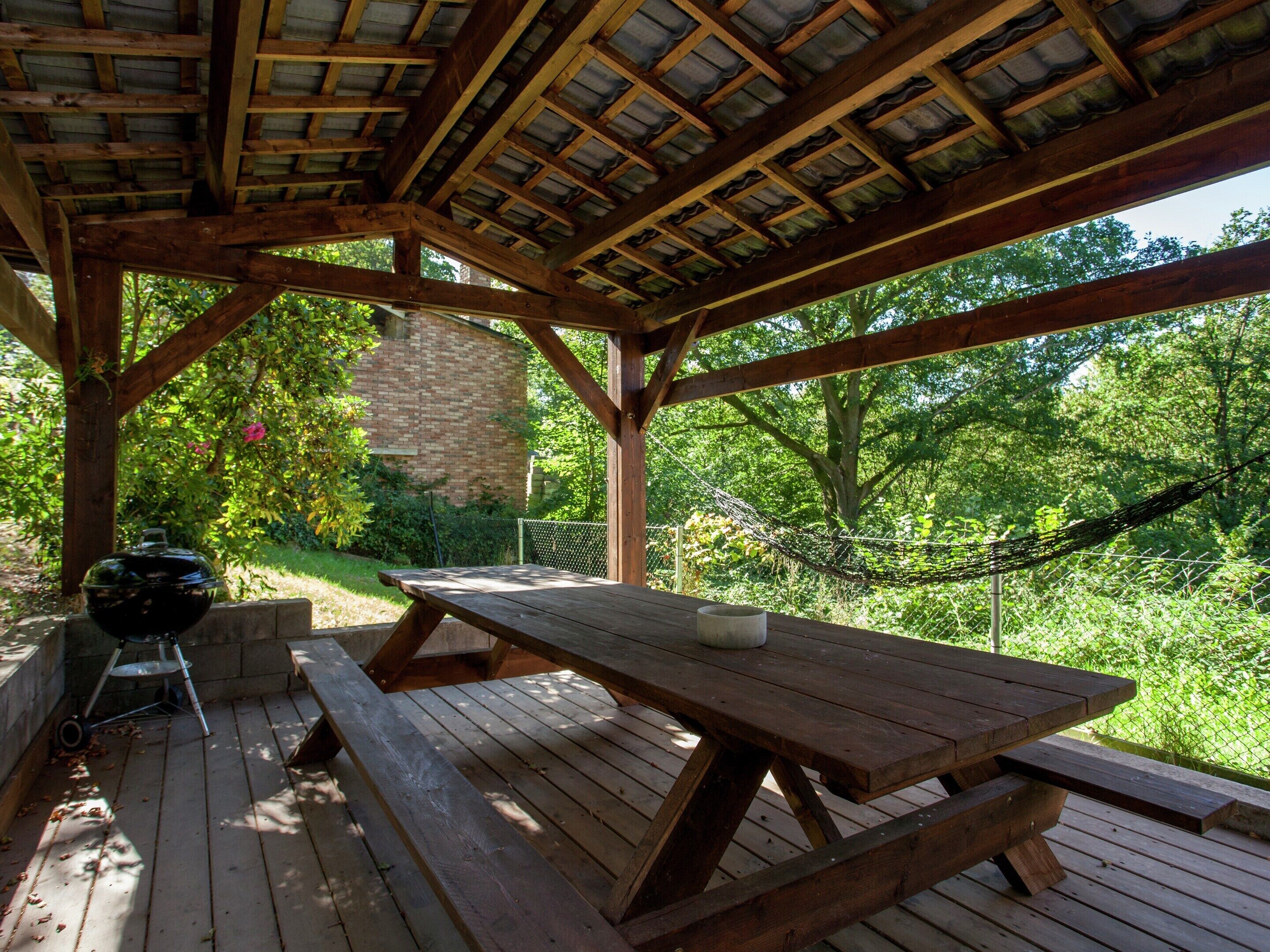 Una terraza de madera con mesas de picnic, una parrilla y un muro de piedra al fondo.