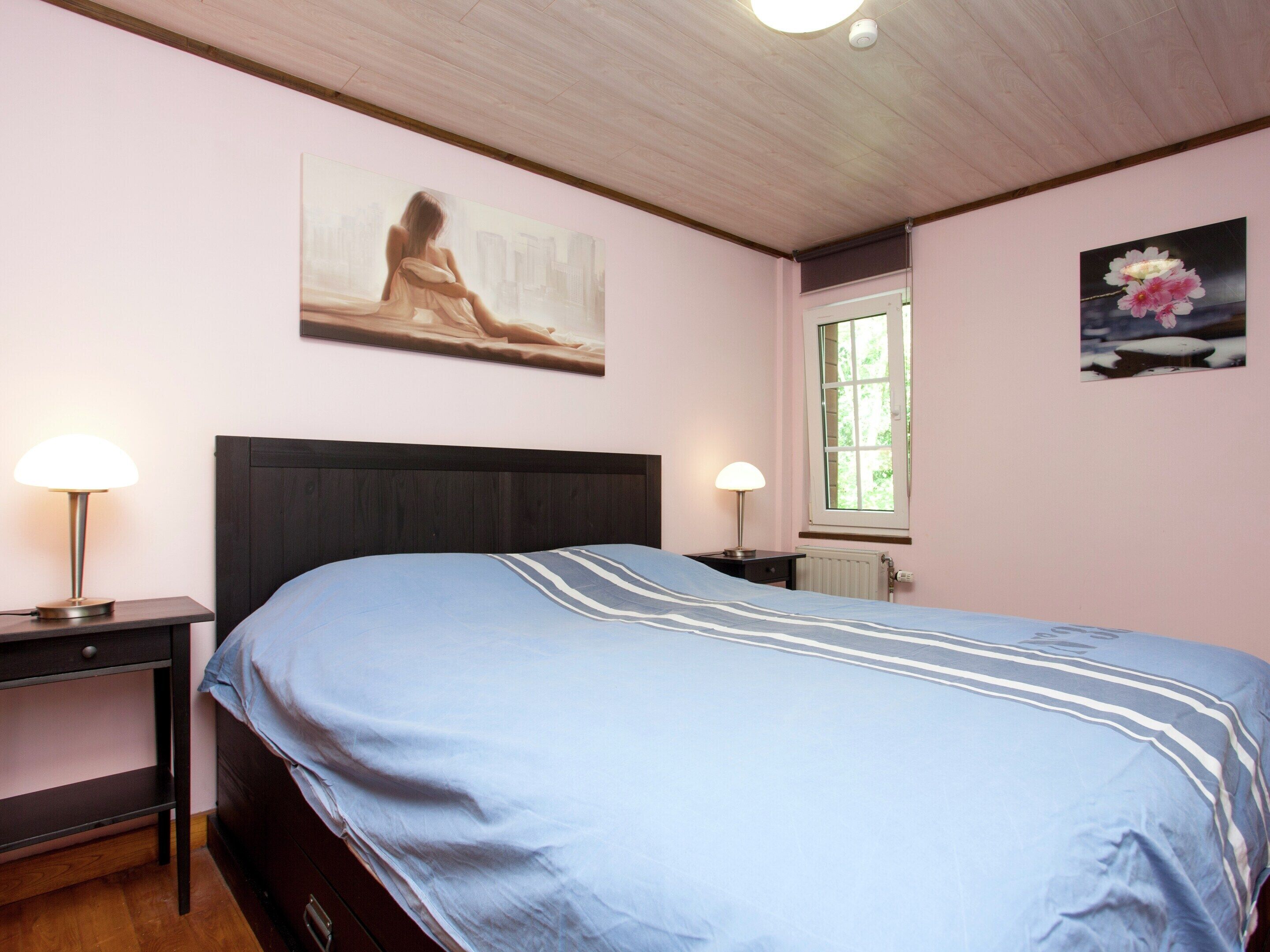 A bedroom with a bed, two bedside lamps, a window, and two wall-mounted pictures.