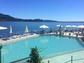 Piscina con sillas de descanso y sombrillas, rodeada por una barandilla y vista a un cuerpo de agua.