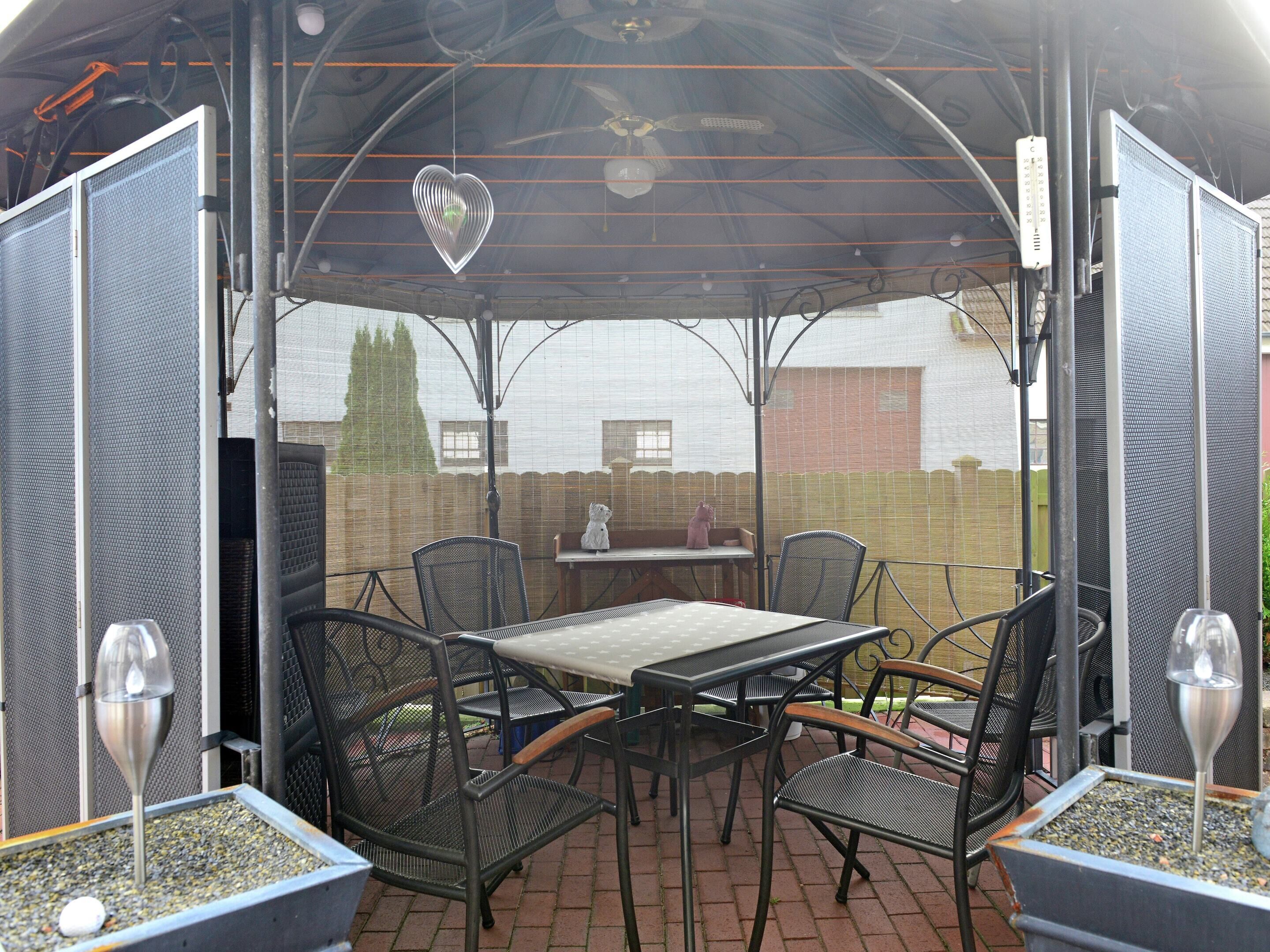 Une terrasse extérieure avec une table et des chaises, un cœur décoratif suspendu au plafond, et une vue sur un mur de briques et des arbres en arrière-plan.