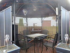 Une terrasse extérieure avec une table et des chaises, un cœur décoratif suspendu au plafond, et une vue sur un mur de briques et des arbres en arrière-plan.