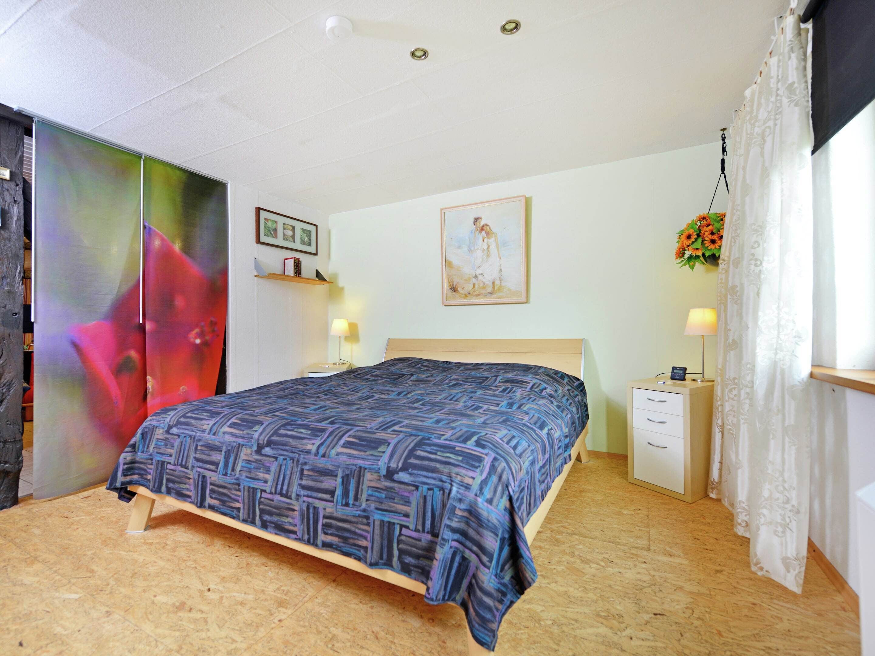 A bedroom with a bed, bedside tables, a nightstand, a wall-mounted shelf, a painting, and a window with curtains.