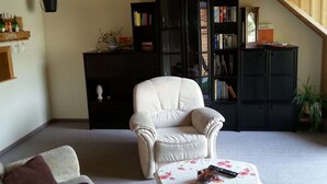 Un salon chaleureux avec un fauteuil blanc, une petite table ronde recouverte d’une nappe à motifs floraux et une bibliothèque remplie de livres.