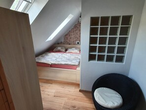 A bedroom with a sloped ceiling, a brick wall, a bed with patterned bedding, a window with multiple panes, and a wicker chair. - Apartment in Traben-trarbach With Balcony (Traben-Trarbach)