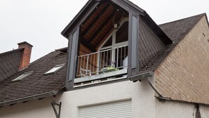 Un immeuble résidentiel doté d’un toit en pente, d’un balcon avec une rambarde en métal et d’une fenêtre avec un volet blanc.