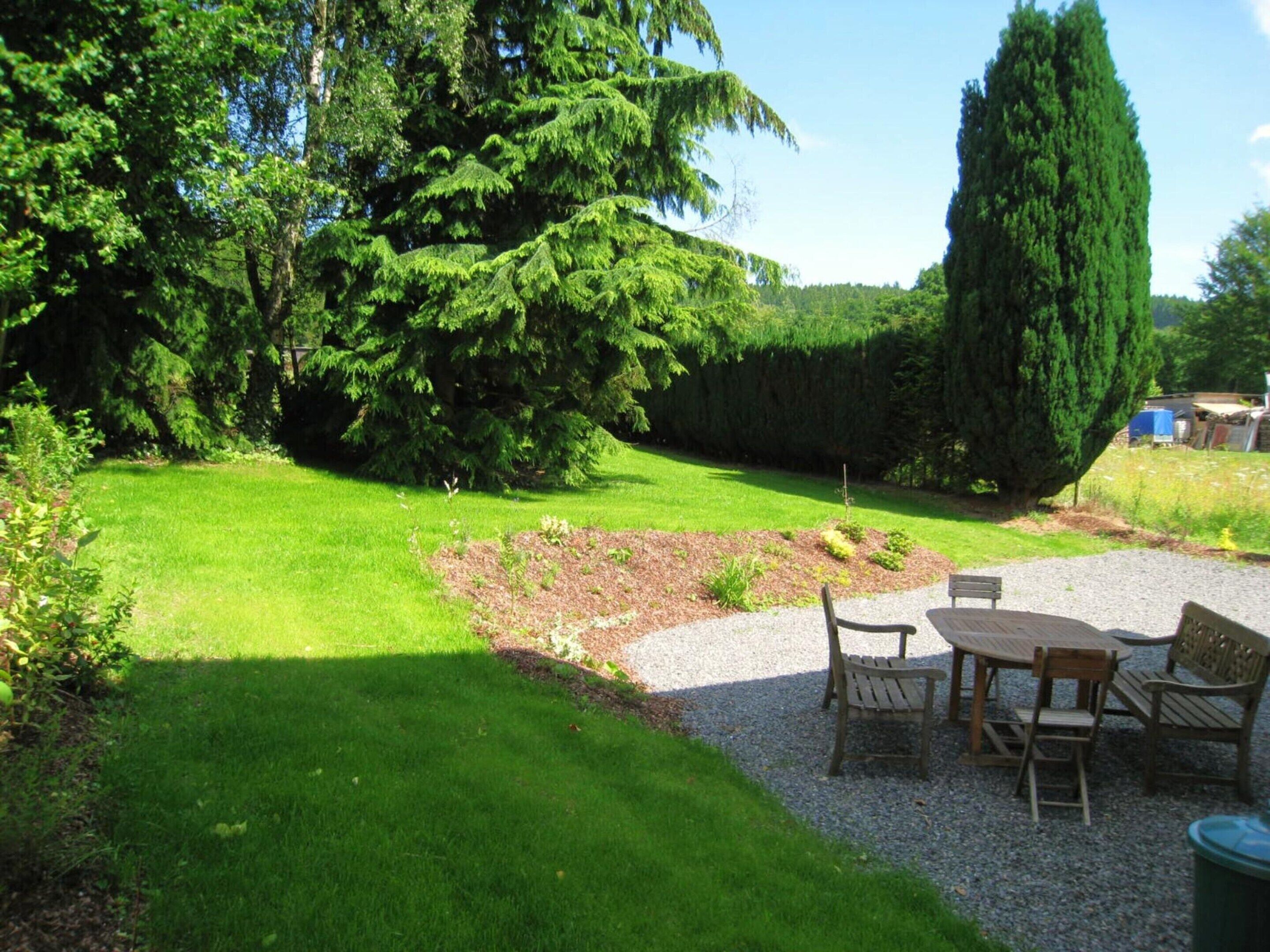 Un giardino curato con un patio in ghiaia, mobili in legno e diverse piante e alberi.