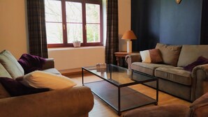 A living room with a sofa, armchair, and a coffee table. There is a window with curtains, a lamp on a side table, and a mounted animal skull on the wall. - Rustic Ardennes Retreat (Vielsalm)