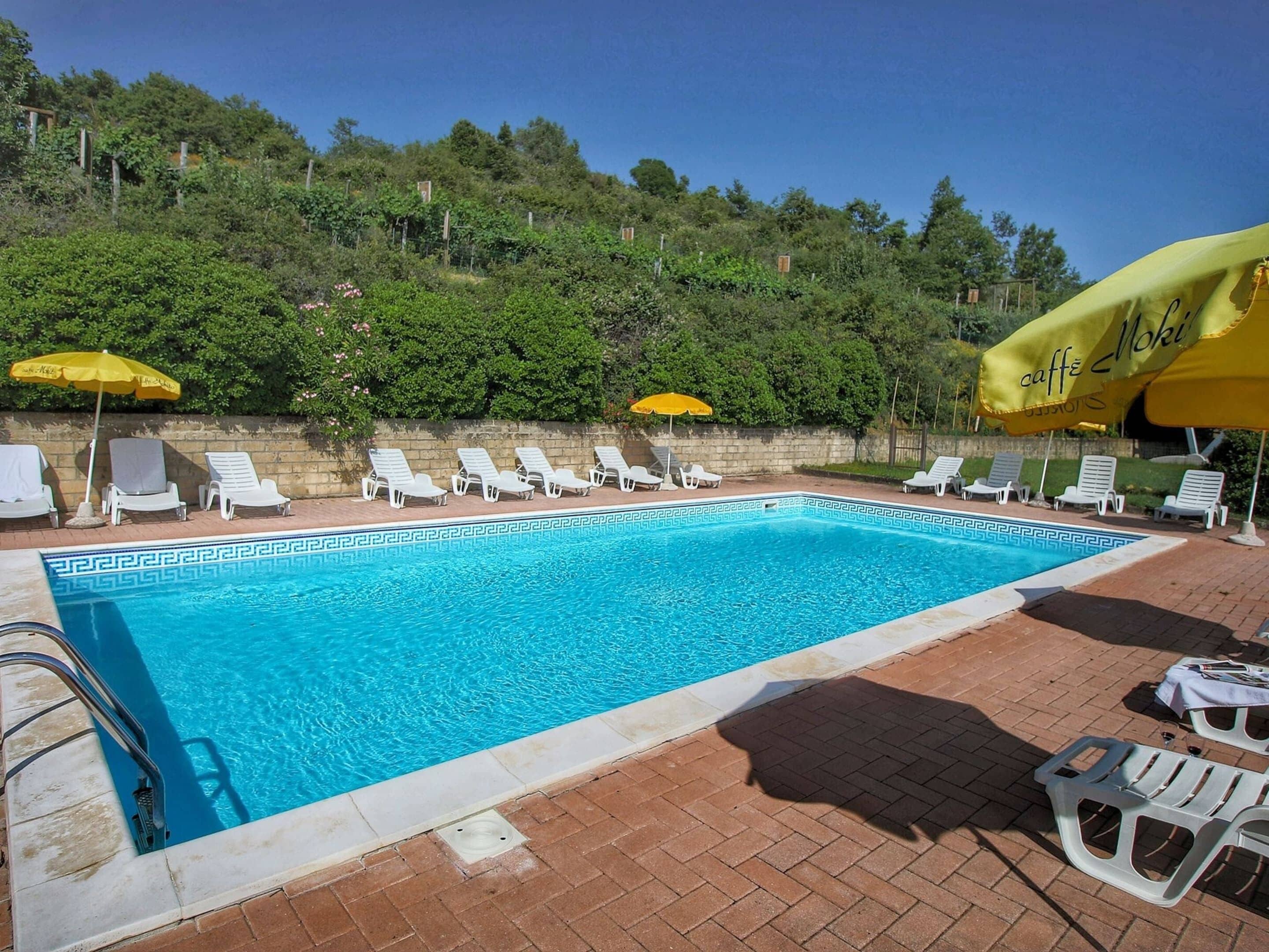 A rectangular swimming pool with a geometric tile pattern, surrounded by white lounge chairs and yellow umbrellas.