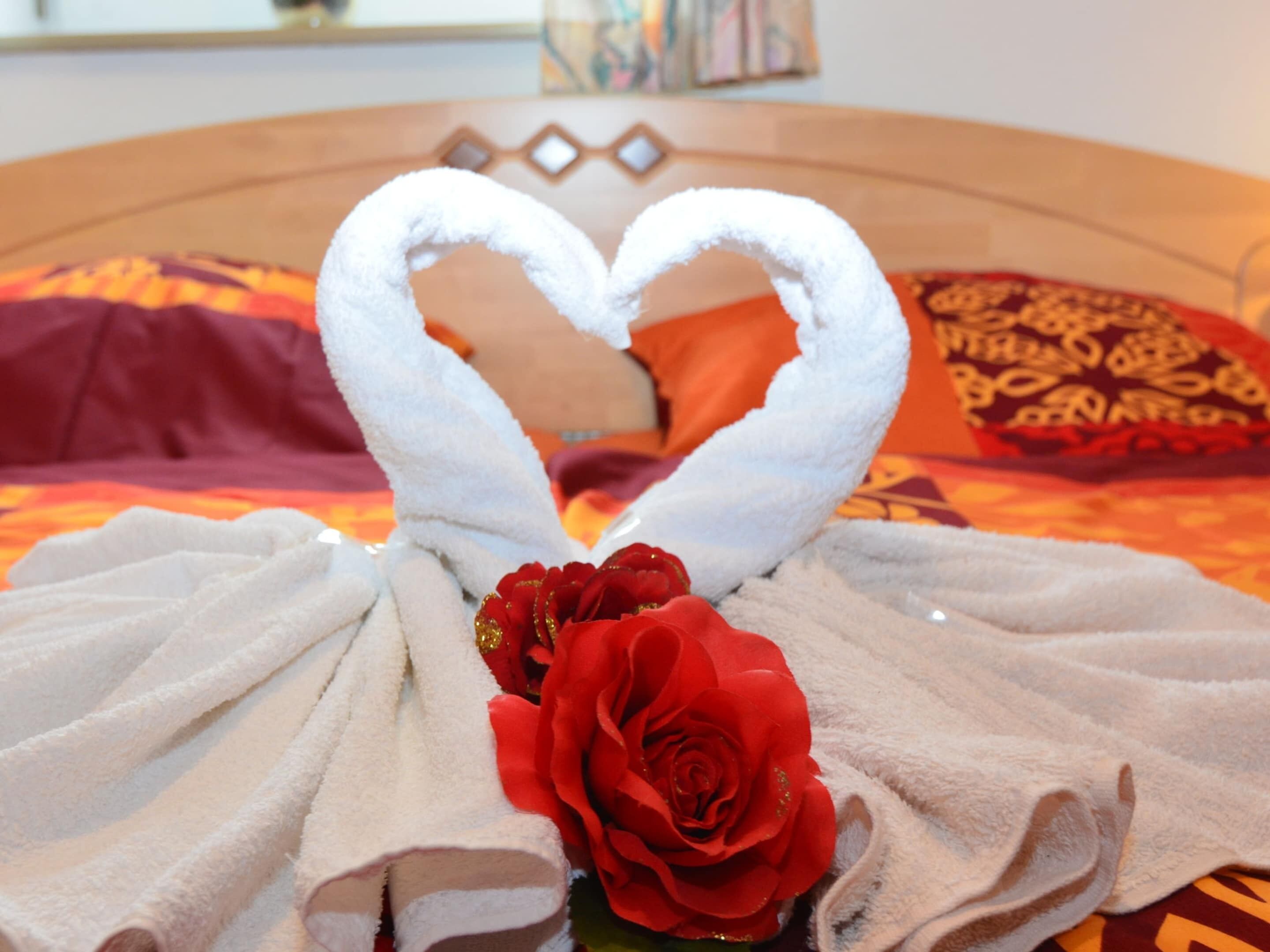 Una cama con una toalla dispuesta en forma de corazón y una rosa roja.