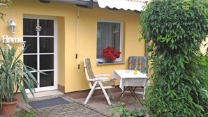 Una casa gialla con una porta bianca, una finestra con fiori rossi, un tavolo bianco e una bottiglia gialla, e una sedia.