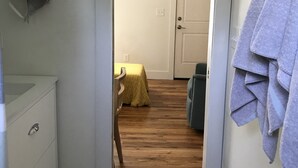 Un couloir étroit mène à une chambre dotée d’un plancher en bois, d’un lit recouvert d’une couvre-lit jaune et d’une porte.