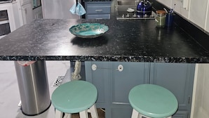 Een keuken met een zwart aanrecht, een roestvrijstalen koelkast, een magnetron en een spoelbak. Er staan twee groene krukken en een decoratieve schaal op het aanrecht.