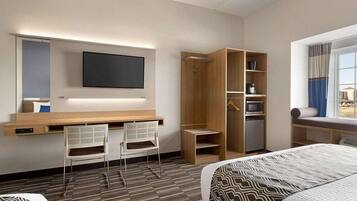 A hotel room with a bed, a desk with two chairs, a TV mounted on the wall, a wardrobe, and a window with curtains.
