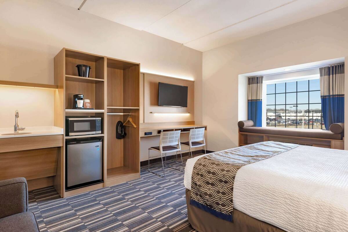 A hotel room with a bed, a desk with a computer, a microwave, a mini-fridge, and a TV.