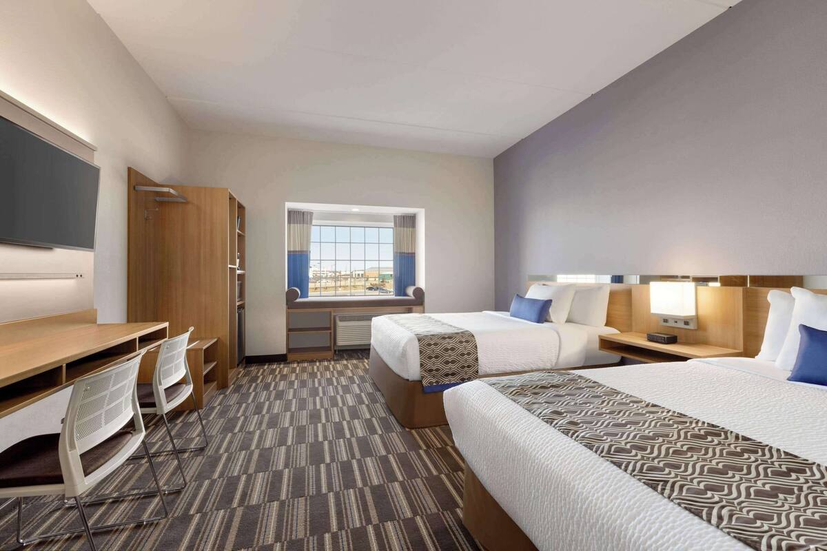 A hotel room with two beds, a desk with chairs, a window with curtains, and a view of the city.