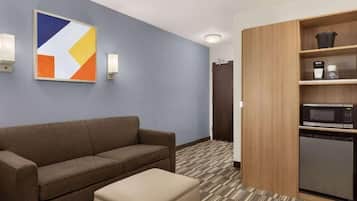 A hotel room with a brown sofa, a beige ottoman, a microwave, a coffee maker, and a cabinet with shelves.