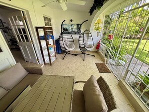 A room with a ceiling fan, a dining table with chairs, a sofa, a bookshelf, and a view of a garden through a window with a metal gate.