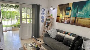 A living room with a glass-top coffee table, a grey sofa with decorative pillows, a wall-mounted fan, and a bookshelf with books and decorative items. - West Coast 1 Bedroom Apartment - Close to beach (66) (Holetown)