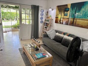 A living room with a glass-top coffee table, a grey sofa with decorative pillows, a wall-mounted fan, and a bookshelf with books and decorative items. - West Coast 1 Bedroom Apartment - Close to beach (66) (Holetown)