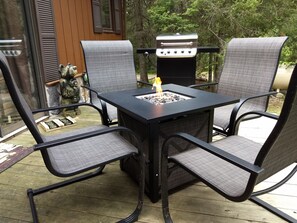 Un patio con una mesa de metal negro y cuatro sillas de malla gris, una parrilla de carbón y un brasero con fuego encendido.