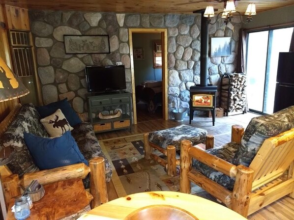 Interior rústico de una cabaña con pared de piedra, mobiliario de madera, chimenea y televisor.