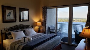 Ein Hotelzimmer mit einem Bett, zwei gerahmten Bildern, einem Schreibtisch mit Lampe und einem Balkon mit Stuhl.