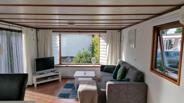 Una sala de estar acogedora con un sofá, televisión y vistas a un jardín a través de amplias ventanas.