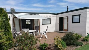 Una casa móvil blanca con un área de patio que incluye una mesa y sillas, rodeada de diversas plantas y arbustos.